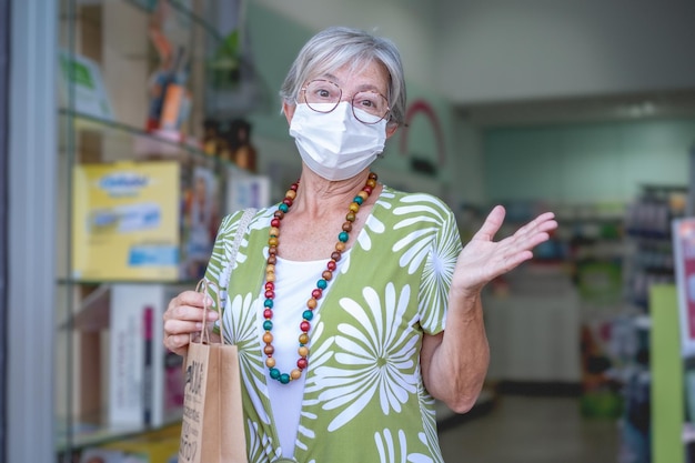 Porträt einer älteren Frau mit Papiertüte, die nach dem Einkauf medizinischer Produkte in der Apotheke eine Maske trägt Hübsche ältere Dame mit Brille in der Drogerie-Apotheke