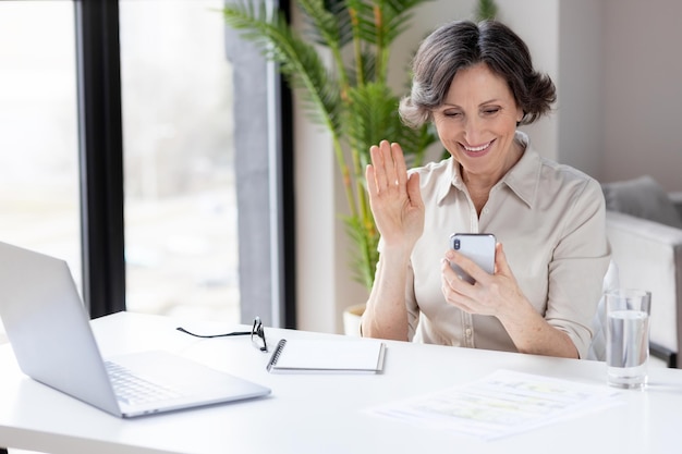 Porträt einer älteren Geschäftsfrau, die an einem Büro- oder Heimschreibtisch sitzt, verwendet ein Mobiltelefon, um über das Internet zu kommunizieren. Technologie, Videoanruf, Online-Konferenzkonzept