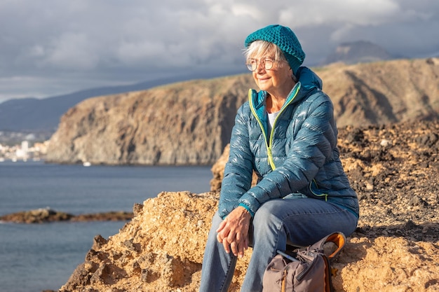Porträt einer älteren lächelnden Frau, die sich bei einem Ausflug im Freien auf See ausruht und auf der Klippe mit Blick auf den Horizont sitzt Fit reife attraktive Frau auf einer Wanderreise, die Freiheit und gesunden Urlaub genießt