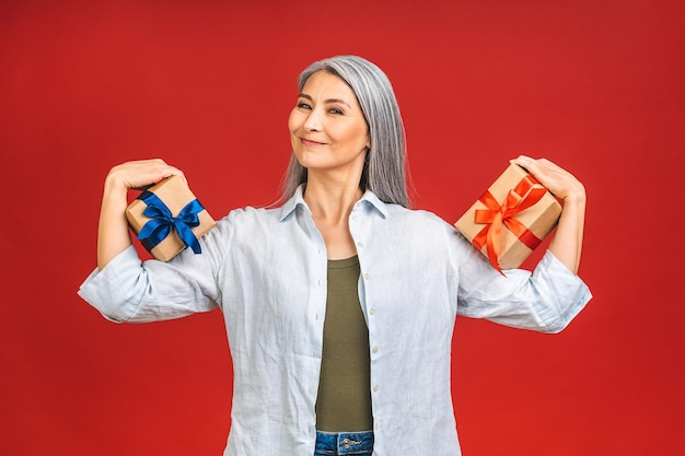 Porträt einer älteren, reifen, schönen Dame, die Geburtstagsgeschenk-Geschenkboxen über isoliertem rotem Hintergrund hält und mit glücklichem Lächeln und Siegerausdruck die Leistung feiert