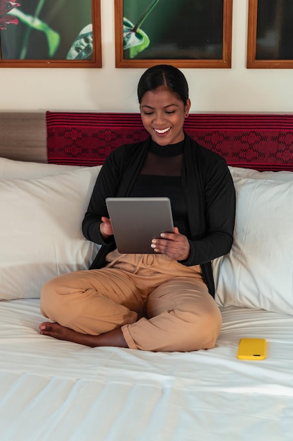 Porträt einer afroamerikanischen Frau, die im Bett sitzt und ihr digitales Tablet überprüft.