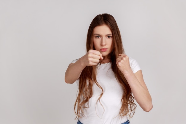 Porträt einer aggressiven Frau, die geballte Fäuste hochhält und mit Täterverteidigung vor häuslicher Gewalt kämpft und weißes T-Shirt trägt. Studioaufnahme isoliert auf grauem Hintergrund