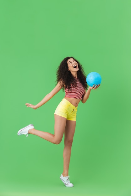 Porträt einer aktiven Frau der 20er Jahre, die Sommerkleidung trägt und lächelt und Volleyball hält, während sie auf Grün steht