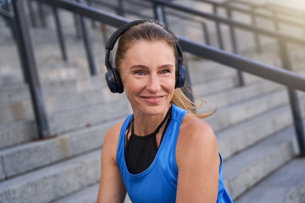 Porträt einer aktiven Frau mittleren Alters in Sportkleidung und Kopfhörern, die in die Kamera lächelt und im Freien posiert