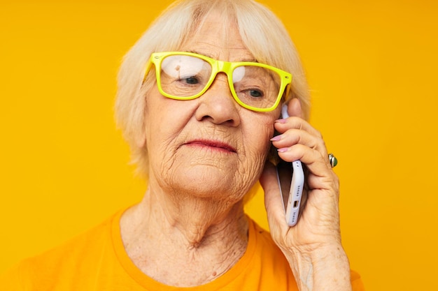 Porträt einer alten freundlichen Frau, die am Telefon in gelber Brille spricht, isolierter Hintergrund