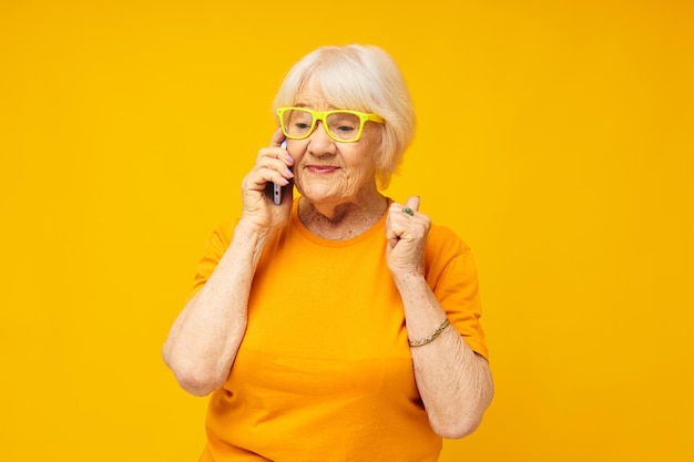 Porträt einer alten freundlichen Frau, die am Telefon in gelber Brille spricht, isolierter Hintergrund