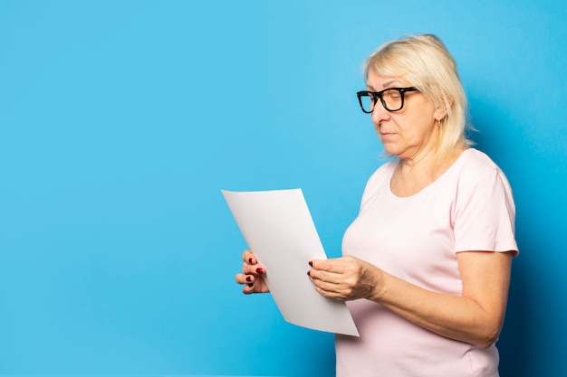 Porträt einer alten freundlichen Frau in einem lässigen T-Shirt und einer Brille, die ein Blatt Papier in ihren Händen halten und es auf einer isolierten blauen Wand betrachten. Emotionales Gesicht. Konzeptbrief, Hinweis