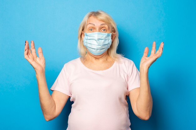 Porträt einer alten freundlichen Frau in einem T-Shirt und einer medizinischen Schutzmaske zuckt die Achseln an der blauen Wand. Emotionales Gesicht. Konzeptvirus, Quarantäne, schmutzige Luft, Pandemie. Geste der Angst, Sorge