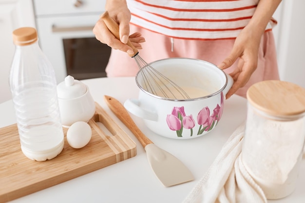 Porträt einer anonymen unbekannten Frau in gestreifter Kleidung, die Teig mit einem Schneebesen in einem Topf mischt, der neben einem Tisch mit Produkten zum Backen in der Küche steht