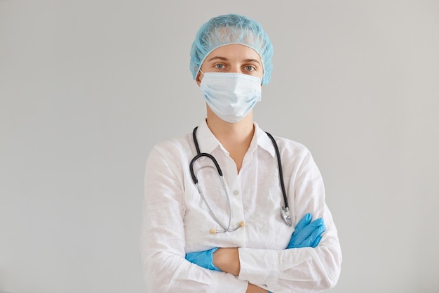 Porträt einer Arztfrau mit Handschuhen, medizinischer Maske und Mütze, Kleid, die mit verschränkten Armen drinnen posiert, selbstbewussten Gesichtsausdruck hat und auf Patienten wartet.