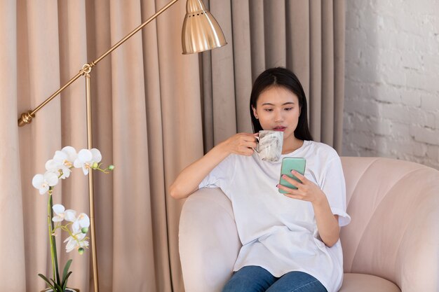 Porträt einer asiatischen Frau, die Nachmittagstee genießt und Nachrichten auf dem Handy im Innenwohnzimmer der Wohnung liest?