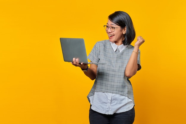 Porträt einer asiatischen Frau mit Brille aufgeregt beim Öffnen eingehender E-Mails auf dem Laptop isoliert auf gelbem Hintergrund