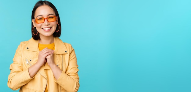 Porträt einer asiatischen Frau mit Sonnenbrille, die hoffnungsvoll aussieht, geschmeichelt lächelnd glücklich über blauem Hintergrund steht