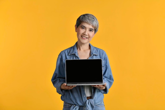 Porträt einer asiatischen Frau mittleren Alters in den 50er Jahren, die ein lässiges Jeanshemd in einem weißen T-Shirt trägt und einen Laptop isoliert auf gelbem Hintergrund hält, der in die Kamera schaut und lächelt