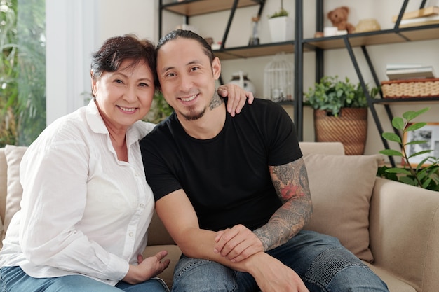 Porträt einer asiatischen glücklichen Mutter, die ihren älteren Sohn umarmt, während sie auf dem Sofa im Zimmer sitzen