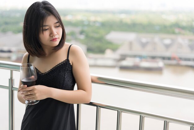 Porträt einer asiatischen jungen hübschen Frau im schwarzen Nachthemd, die auf dem Balkon steht und ein Glas kalten Rotwein hält