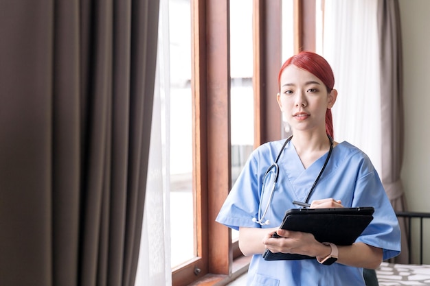 Porträt einer asiatischen Krankenschwester, die ein Stethoskop mit medizinischem Peeling trägt und das Tablet im Haus des Patienten hält