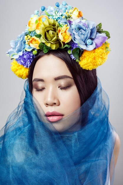 Porträt einer attraktiven asiatischen Frau mit Blumenhut und blauem Schleier, die mit geschlossenen Augen auf hellgrauem Hintergrund posiert. Indoor-Studioaufnahme.