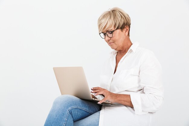 Porträt einer attraktiven erwachsenen Frau mit Brille, die mit Laptop isoliert über weißer Wand im Studio sitzt