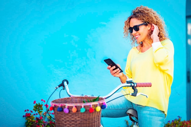 Porträt einer attraktiven erwachsenen Frau mittleren Alters, die am Handy lächelt und auf einem Fahrrad vor einer blauen Wand im Hintergrund sitzt. Frauen genießen die Technologieverbindung im Freien