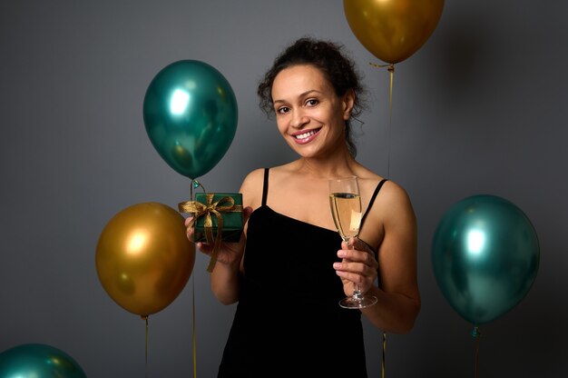 Porträt einer attraktiven jungen Frau im schwarzen Kleid, die ein Glas mit Champagner und ein Weihnachtsgeschenk hält, lächelndes zahniges Lächeln auf einem grauen Wandhintergrund, der mit goldenen und grünen metallischen Luftballons verziert ist