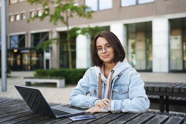 Porträt einer attraktiven jungen Studentin mit Jeansjacke mit kurzen Haaren, die im Freien an einer Bank sitzt und auf einem Tisch sitzt und mit Laptop und Handy arbeitet und zufrieden lächelt und alleine draußen lernt