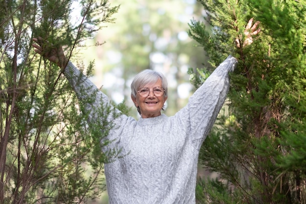 Porträt einer attraktiven kaukasischen weißhaarigen Seniorin mit ausgestreckten Armen im Wald Lächelnde ältere Dame, die im Wald die Freiheit der Bergnatur genießt