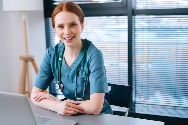 Porträt einer attraktiven lächelnden jungen Ärztin in blaugrüner medizinischer Uniform, die am Schreibtisch mit Laptop auf dem Hintergrund des Fensters sitzt