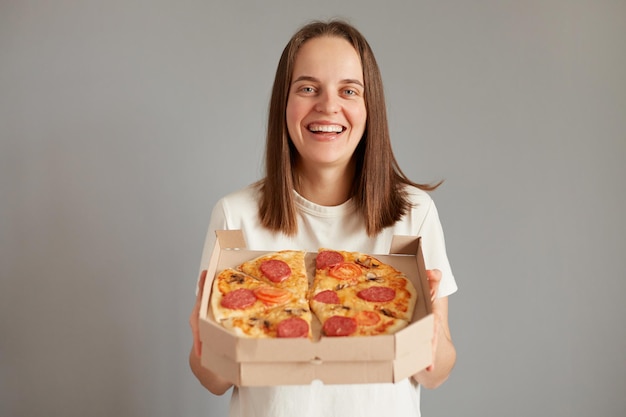 Porträt einer attraktiven, lächelnden, positiv gekleideten, weißen, lässigen T-Shirt-Stehenden, die Pizza zum Mitnehmen isoliert auf grauem Hintergrund hält und Ihnen anbietet, köstliches Fast Food zu probieren