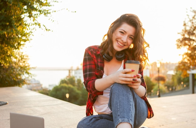 Porträt einer attraktiven lächelnden Studentenfrau. Junges Hippie-Mädchen, das bei Sonnenaufgang eine Tasse Kaffee in der Stadt hält