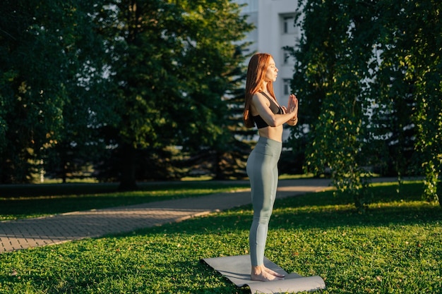 Porträt einer attraktiven rothaarigen jungen Frau mit geschlossenen Augen, die Yoga und Meditation praktiziert und Namaste-Pose durchführt Ruhige Frau, die abends bei Sonnenuntergang im Stadtpark Namaste-Geste macht