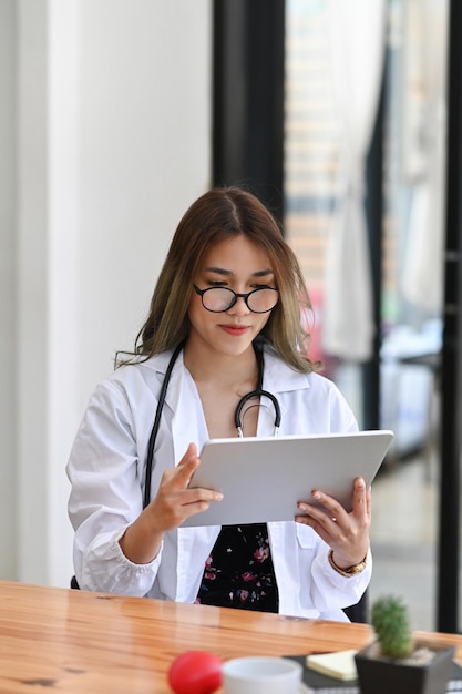 Porträt einer attraktiven Ärztin in weißer Uniform mit digitalem Tablet, während sie in der Arztpraxis sitzt