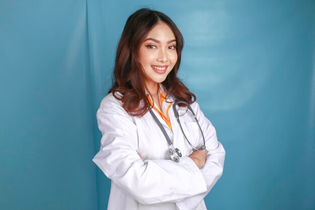 Porträt einer attraktiven, selbstbewussten Ärztin, die freundlich lächelnde Arme verschränkt trägt, trägt ein weißes Laborkittel-Stethoskop, isoliert auf blauem Hintergrund