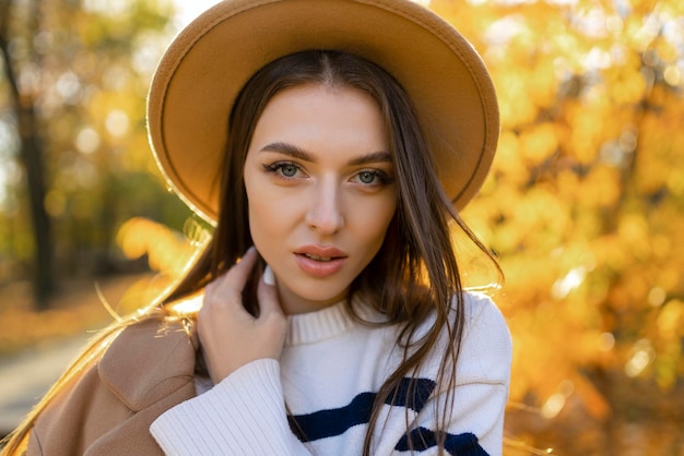 Porträt einer attraktiven, stilvollen, lächelnden Frau mit langen Haaren, die im Park spazieren geht, gekleidet in einem warmen, braunen Mantel, Herbst, trendiger Mode-Streetstyle mit Hut