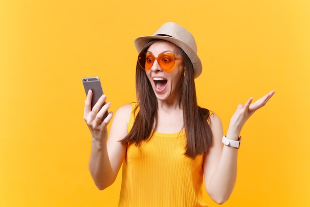 Porträt einer aufgeregten, erstaunten jungen Frau mit Strohsommerhut, orangefarbener Brille, die den Mund weit offen hält, überrascht aussieht, das Handy isoliert auf gelbem Hintergrund hält. Konzept der aufrichtigen Emotionen der Menschen.