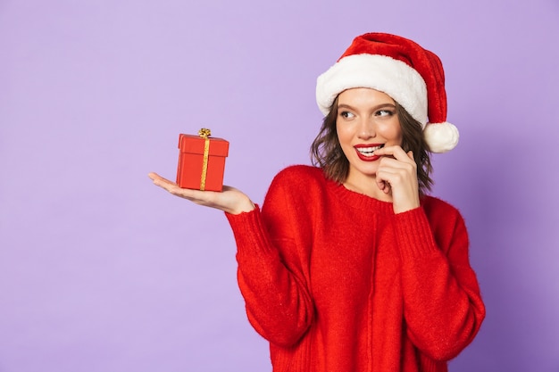 Porträt einer aufgeregten glücklichen jungen Frau, die Weihnachtshut lokalisiert über lila Wand hält, die Überraschungsgeschenkbox hält.