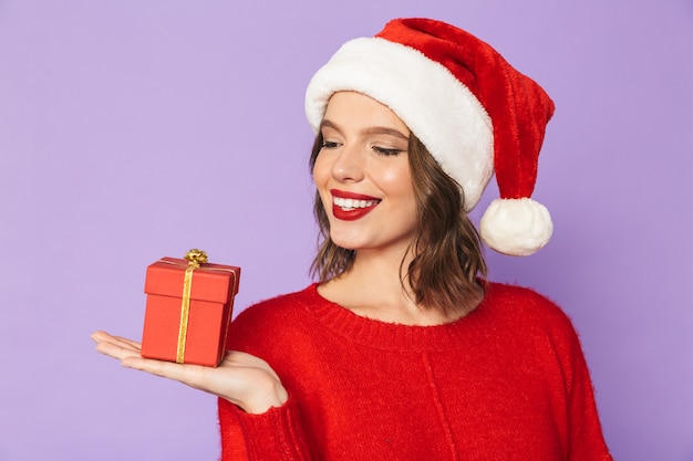 Porträt einer aufgeregten glücklichen jungen Frau, die Weihnachtshut lokalisiert über lila Wand hält, die Überraschungsgeschenkbox hält.