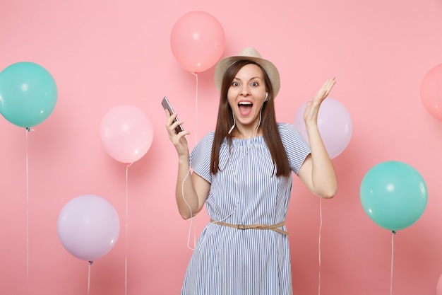 Porträt einer aufgeregten jungen Frau in Strohsommerhut und blauem Kleid mit Handy und Kopfhörern, die Musik hören, die Hand auf rosafarbenem Hintergrund mit bunten Luftballons ausbreitet. Geburtstagsfeier.