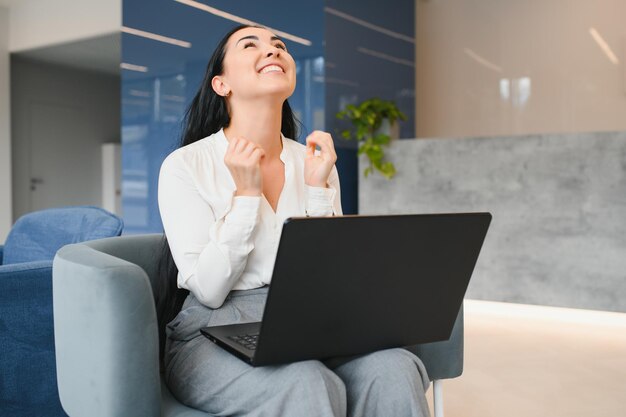 Porträt einer aufgeregten jungen kaukasischen Frau, die Erfolg feiert, während sie zu Hause oder im modernen Büro mit Laptop sitzt Fröhliche Freiberuflerin, die im Coworking Space arbeitet