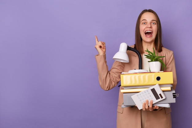 Porträt einer aufgeregten, positiv lachenden, braunhaarigen jungen erwachsenen Frau mit beigefarbener Jacke, die viele Dokumentenordner isoliert über lila Hintergrund hält und auf den Kopierbereich zeigt