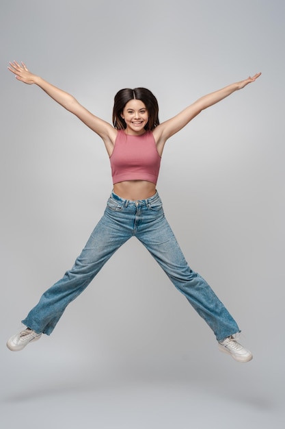 Porträt einer aufgeregten Teenagerin in rosafarbenem Top und Jeans, die isoliert auf grauem Hintergrund springt