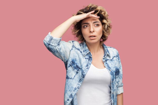 Porträt einer aufmerksamen jungen Frau mit lockiger Frisur im lässigen blauen Hemd, die mit der Hand auf der Stirn steht und mit ernstem Gesicht und Aufmerksamkeit wegschaut. Studioaufnahme, auf rosa Hintergrund isoliert.
