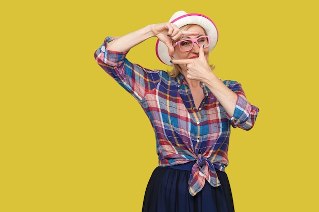 Porträt einer aufmerksamen, modernen, stilvollen, reifen Frau im lässigen Stil mit Hut und Brille, die mit Erntekompositionsgeste steht und das Ziel betrachtet. Indoor-Studioaufnahme isoliert auf gelbem Hintergrund