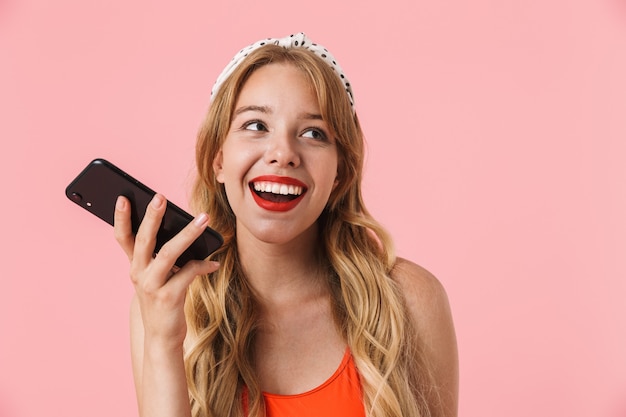 Porträt einer begeisterten jungen Frau mit langen lockigen Haaren, die lächelt und Smartphone isoliert über rosafarbener Wand hält