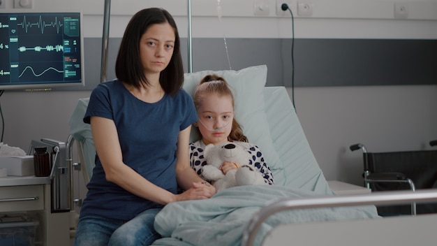 Porträt einer besorgten Mutter, die neben einem im Krankenhaus behandelten kleinen Kind sitzt und in die Kamera schaut