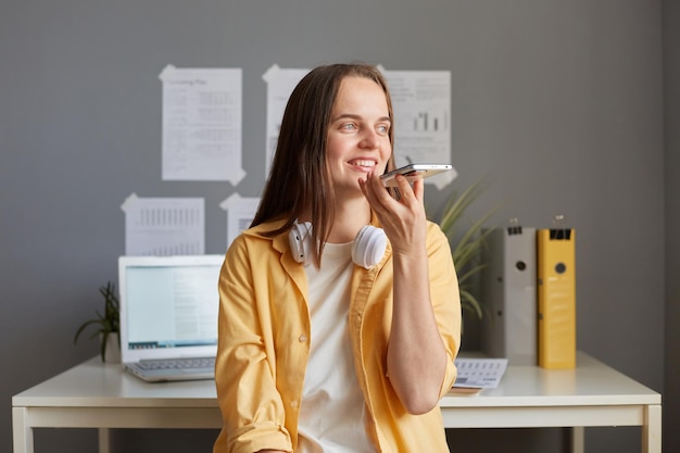 Porträt einer bezaubernden kaukasischen Frau mit gelbem Hemd, die im Büro sitzt und ihr Handy in den Händen hält, um Sprachnachrichten aufzunehmen oder einen Sprachassistenten zu verwenden