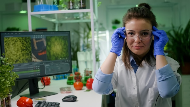 Foto porträt einer biologenfrau, die eine medizinische brille aufsetzt und in die kamera schaut, während sie im pharmazeutischen labor am tisch sitzt. spezialisten, die einen dna-test für genetische mutationen entwickeln