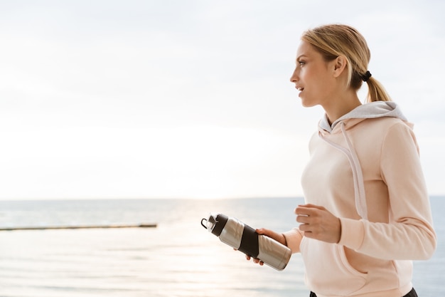 Porträt einer blonden Frau mit Trainingsanzug, die eine Wasserflasche hält, während sie morgens auf dem Pier in der Nähe des Meeres trainiert