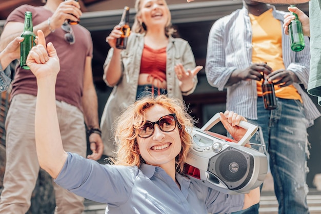 Porträt einer blonden, glücklichen Frau, die eine Stereoanlage hält und auf einer Party um Freunde tanzt