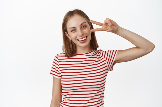 Porträt einer blonden positiven Frau, eines Mädchens, das ein Peace-V-Zeichen zeigt und breit mit weißen, perfekten Zähnen lächelt, glücklich im sommerlich gestreiften T-Shirt vor weißem Hintergrund